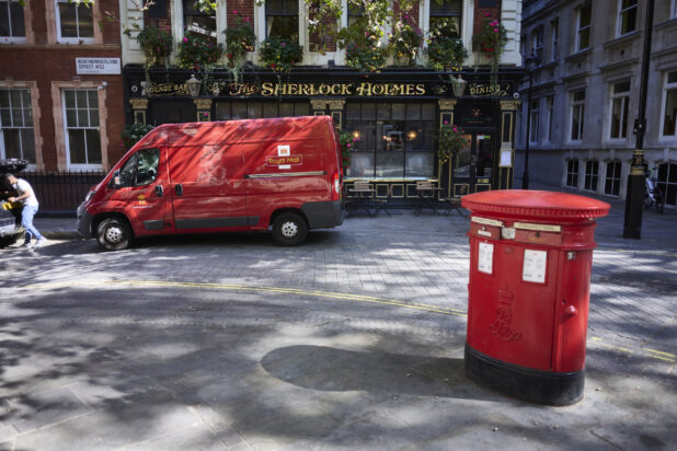 Street view of red post box, mailbox on the edge of the street with a red postal truck in front of a pub in the background, London, England