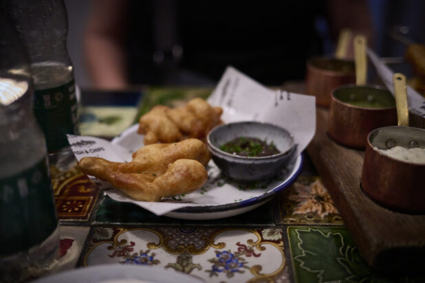 Traditional fish and chips in a pub setting with various sauces in individual copper pots on a wooden board, 2 bottles of water on the side