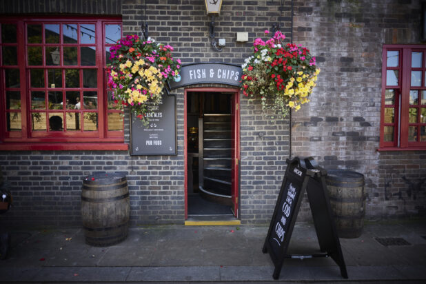 Fish and chip restaurant facade with a sandwich board, wooden barrels, hanging flowers and red windows