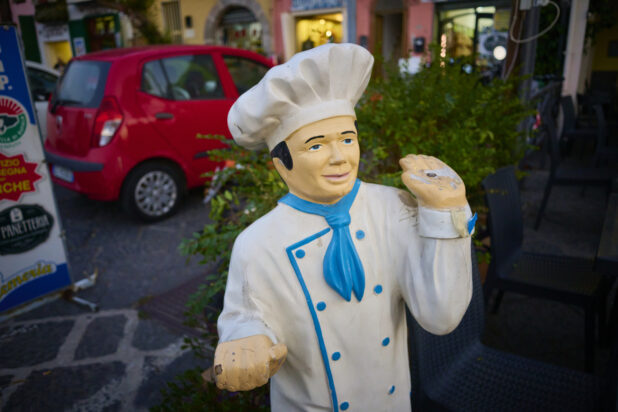 Picture of a chef statue with flowers, a red car and restaurants in the background