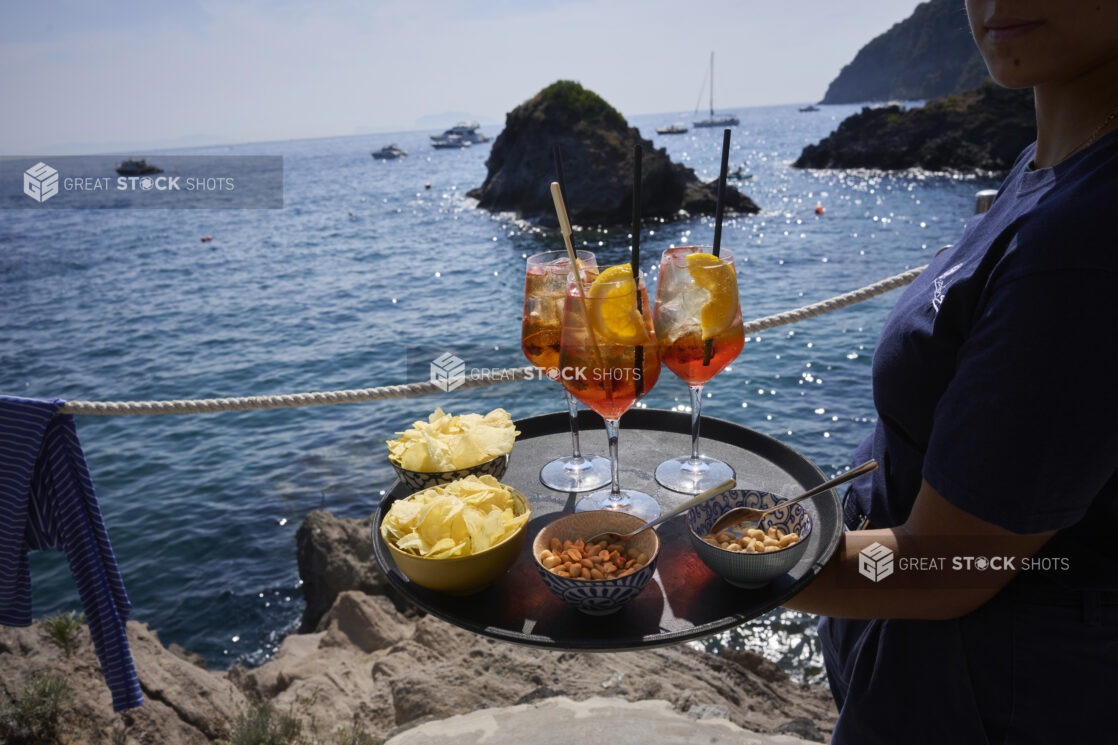 Three Aperol Spritz Cocktails on a tray with Midday Snacks of Chips and nuts being carried out oceanside by a server