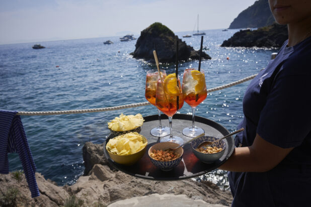 Three Aperol Spritz Cocktails on a tray with Midday Snacks of Chips and nuts being carried out oceanside by a server