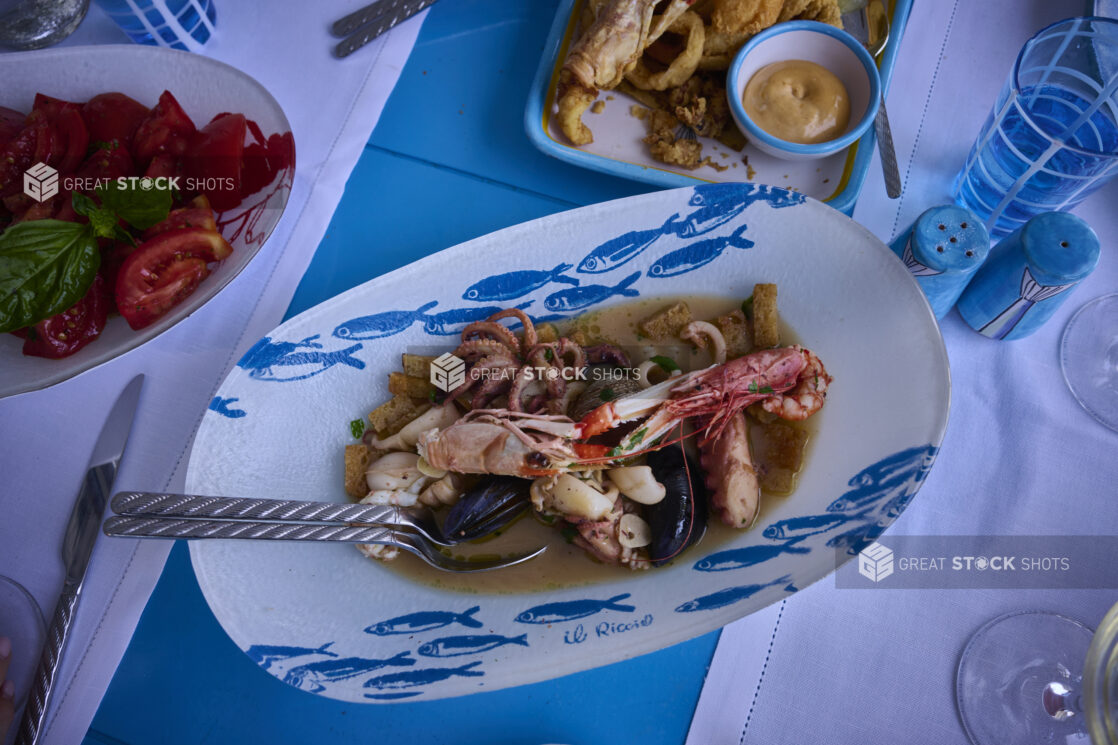 Overhead view of a Mediterranean style meal with a fresh seafood platter, fried seafood platter, fresh tomatoes and basil with seafood patterned dishes, bright blue accessories