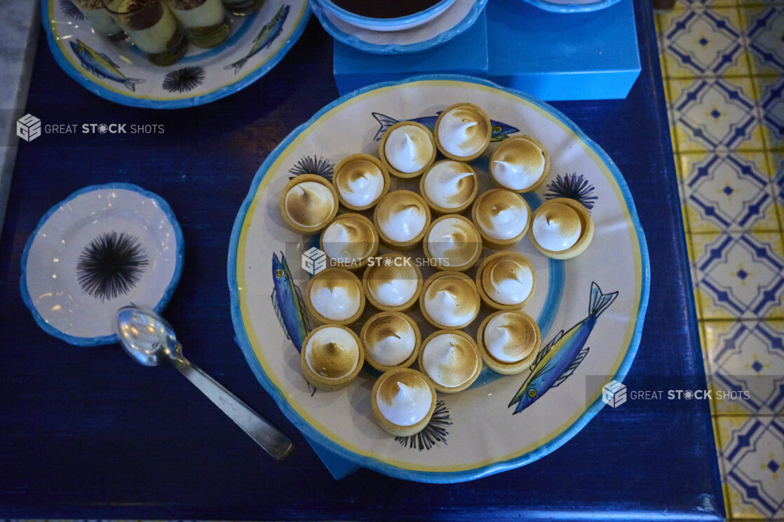 Overhead view of a platter of mini lemon meringue tarts on a table with bright blue accents and Italian tiles