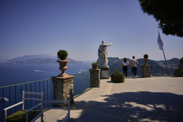 2 people standing beside a statue of Caesar Augustus overlooking the Mediterranean Sea from a cliffside in Capri, Italy, hills and mountains in the background