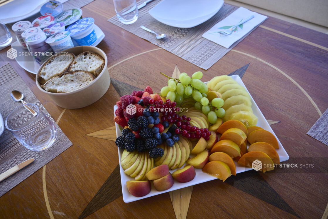 Artful and Colourful Fresh Fruit Tray on a white platter with Fresh Bread and Greek Yogurt and a Place Setting in the Background