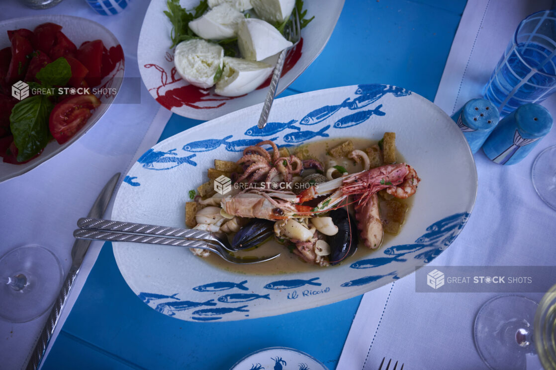 Overhead view of a Mediterranean style meal with a fresh seafood platter, burrata and fresh tomatoes and basil with seafood patterned dishes, bright blue accessories