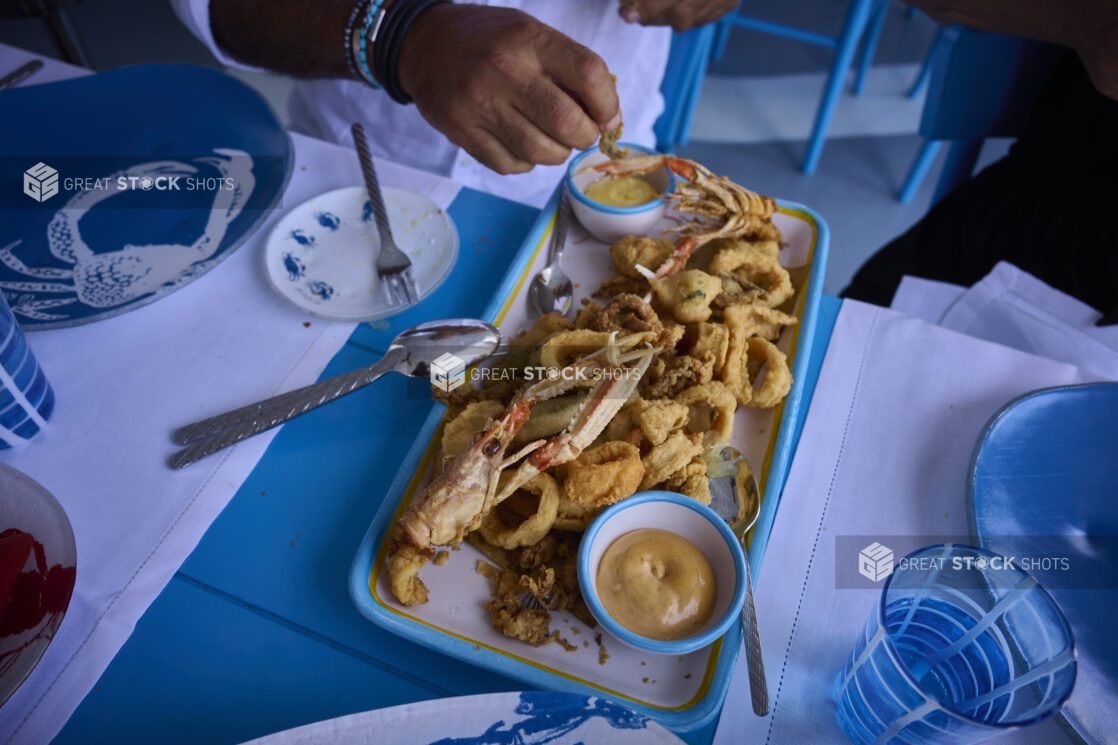 Overhead view of a fried seafood platter with a person eating from the platter with seafood patterned dishes, bright blue