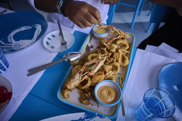 Overhead view of a fried seafood platter with a person eating from the platter with seafood patterned dishes, bright blue