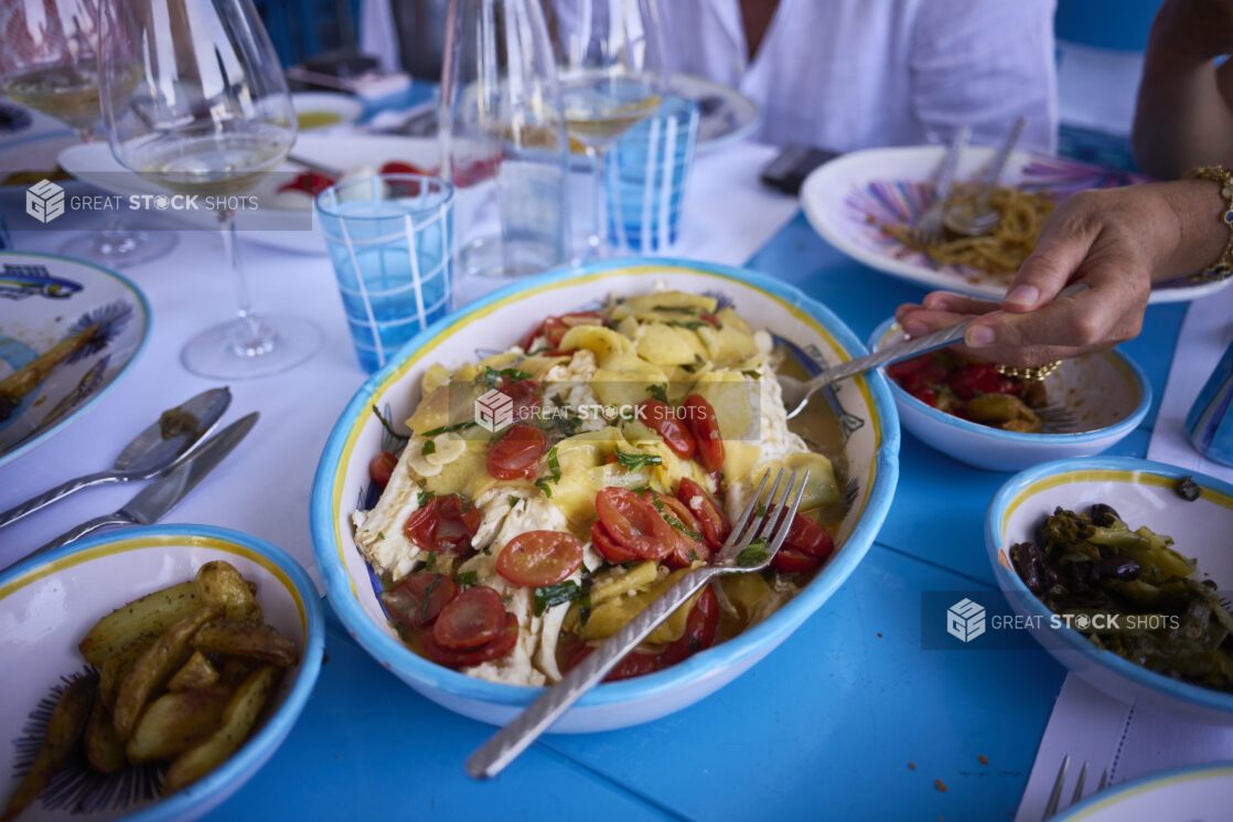 Lunch scene of family style platters of food with people, table settings, bright blue table