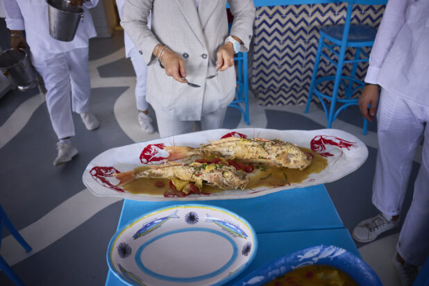 Whole fried fish on a platter being served with bright blue table and chairs, chefs in the background, Mediterranean vibe