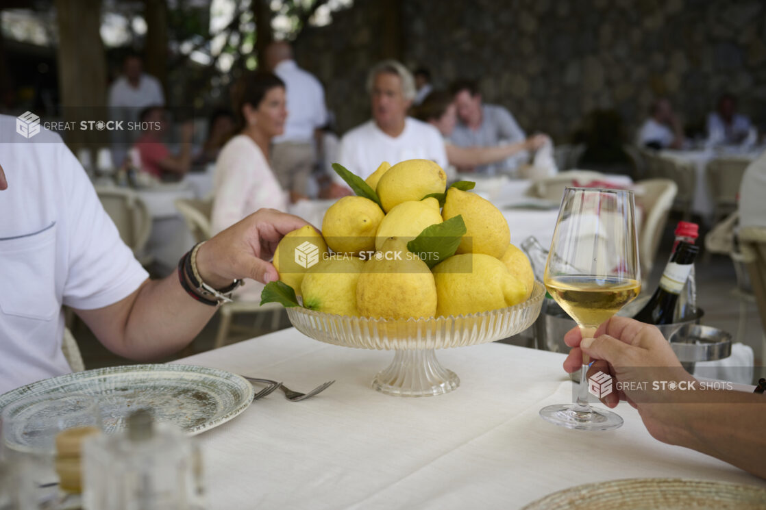 Restaurant scene with a cake stand full of lemons at a table with a hand holding a wine glass and another hand grabbing a lemon