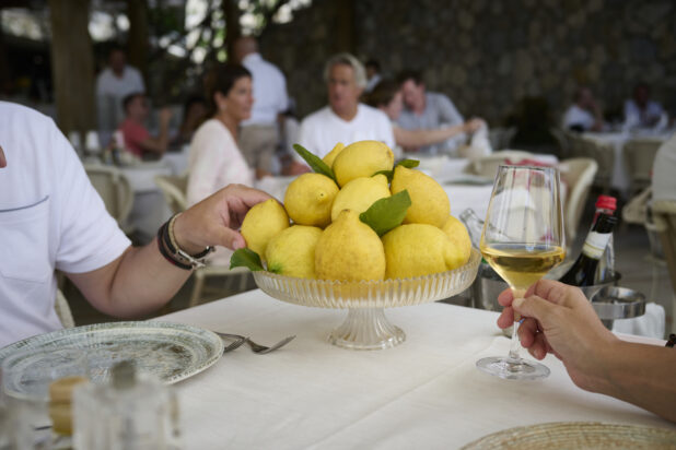 Restaurant scene with a cake stand full of lemons at a table with a hand holding a wine glass and another hand grabbing a lemon