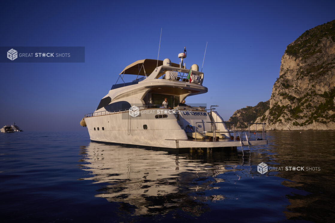 View of a yacht with people, on the sea with a hillside in the background and other yachtws