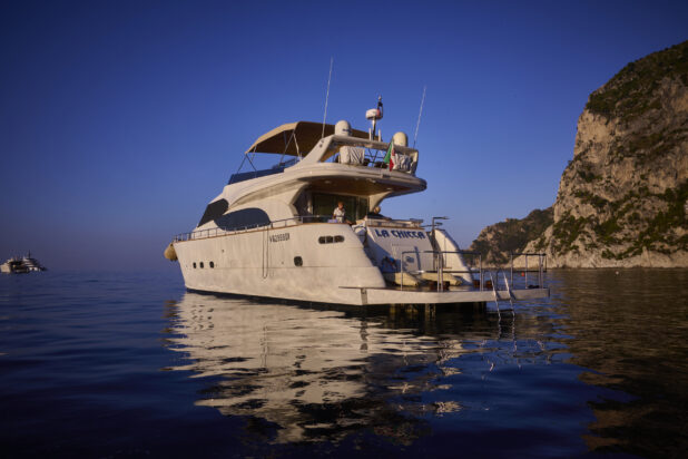 View of a yacht with people, on the sea with a hillside in the background and other yachtws