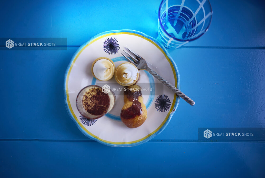 Elegant desserts displayed on a colourful plate with a blue and yellow trim with a fork and a glass of water on a aqua blue background