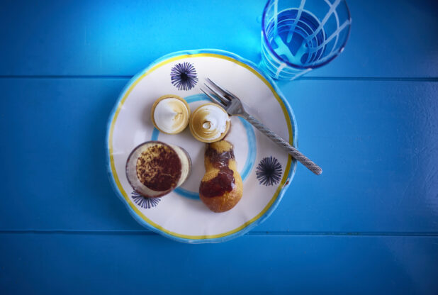 Elegant desserts displayed on a colourful plate with a blue and yellow trim with a fork and a glass of water on a aqua blue background
