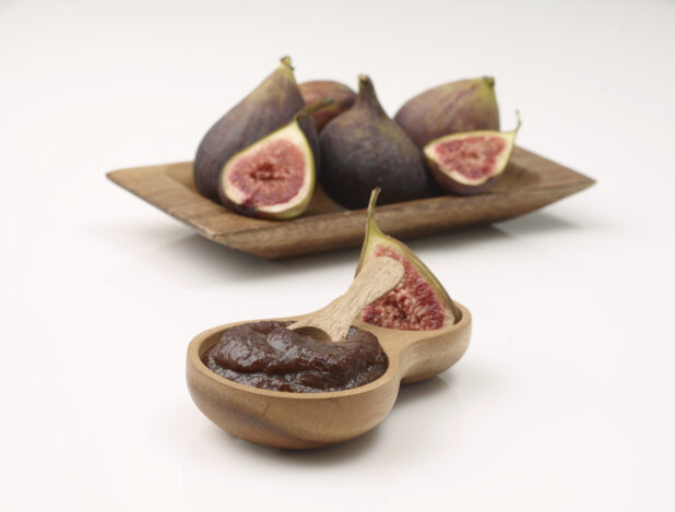 Wooden Bowl of Puréed Fig Sauce with a Wooden Spoon and Fresh Cut and Whole Figs in Background Isolated on a White Background