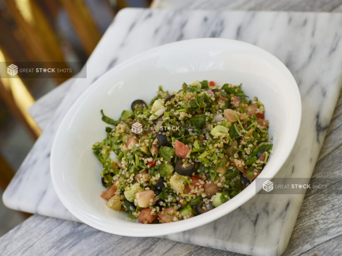 Middle Eastern salad with cous cous, parsley, onion, tomatoes, cucumber and black olives in a white bowl on a marble background