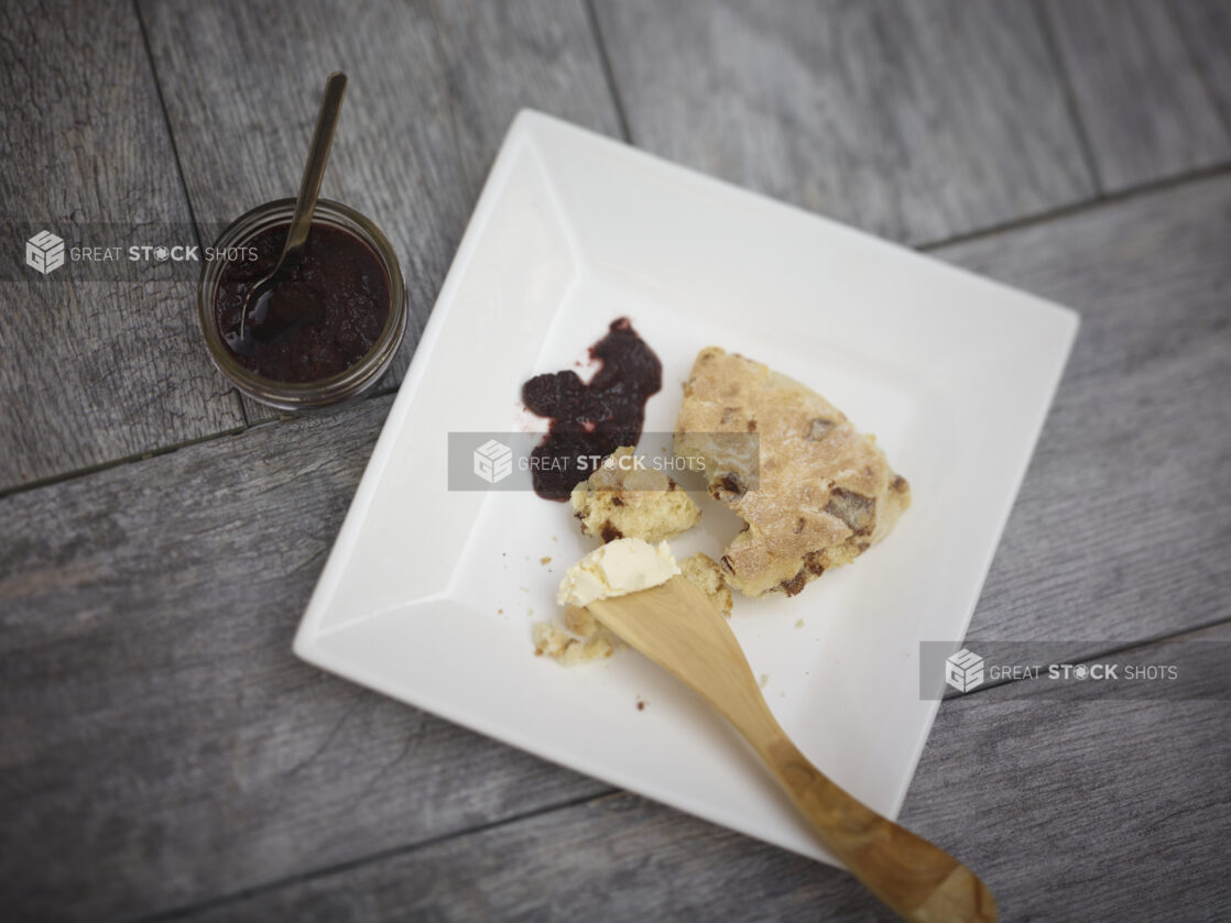 Baked scone/tea biscuit on a square white side plate with a wooden cheese spreader, jam, butter and jam in a small mason jar on a grey wooden background