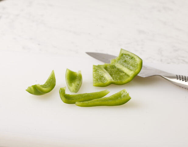 Green bell pepper sliced with a kitchen knife on a white cutting board