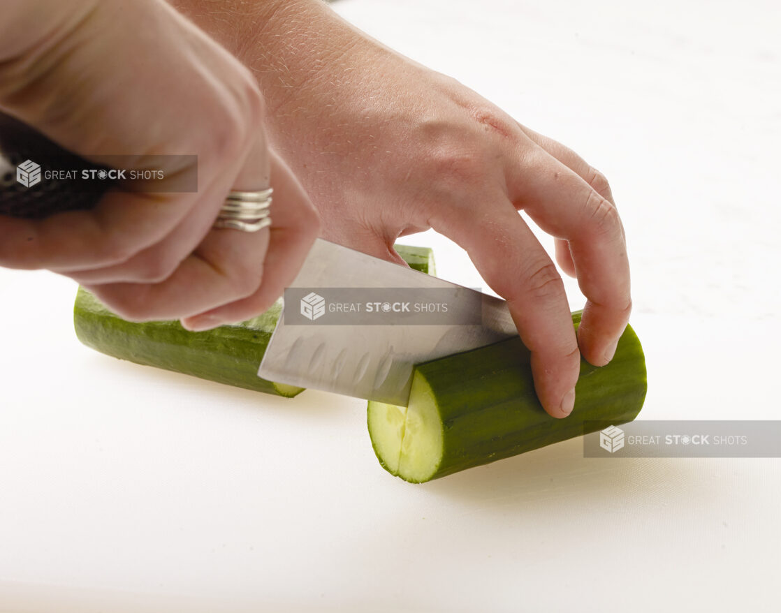 Woman cutting a cucumber in half on a white cutting board with a white marble background