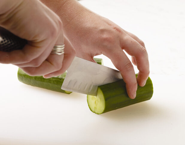 Woman cutting a cucumber in half on a white cutting board with a white marble background