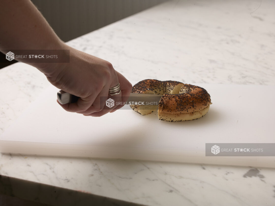 Woman cutting a poppy seed bagel in half on a white cutting board with a white marble background