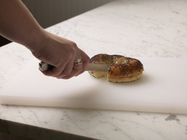 Woman cutting a poppy seed bagel in half on a white cutting board with a white marble background