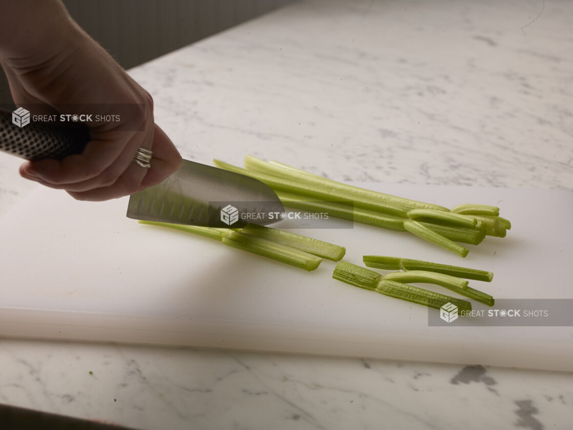 Woman cutting celery sticks on a white cutting board with a white marble background