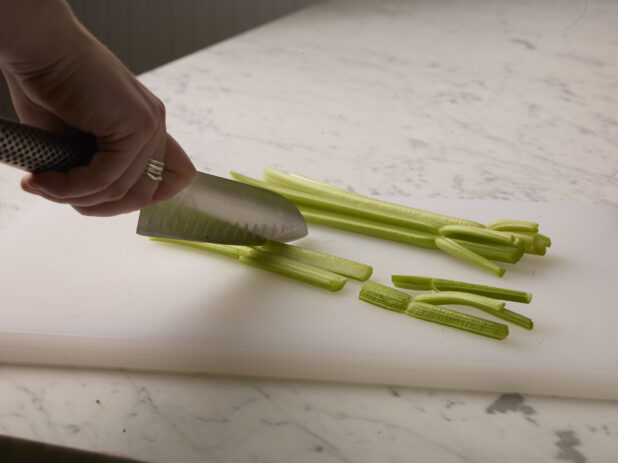 Woman cutting celery sticks on a white cutting board with a white marble background