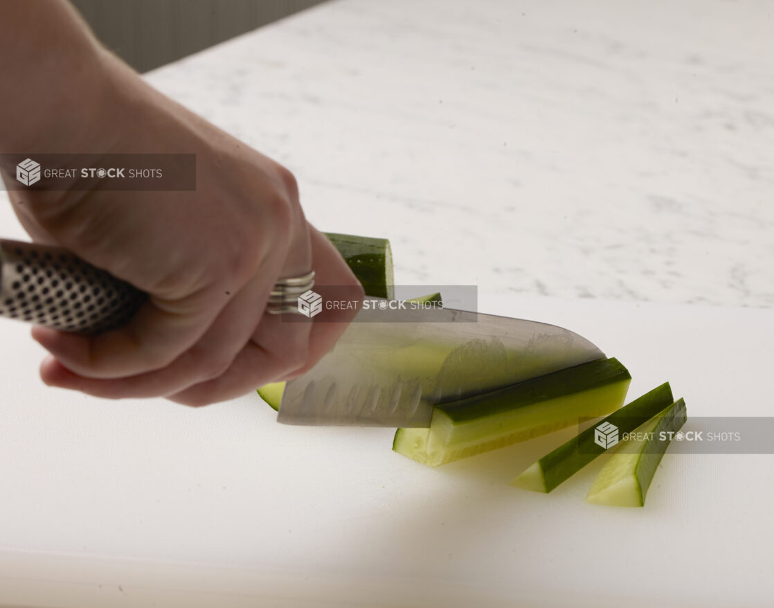 Woman cutting cucumber sticks on a white cutting board with a white marble background