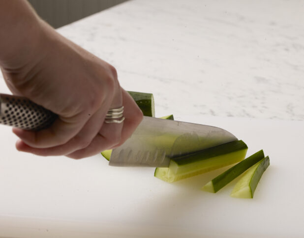 Woman cutting cucumber sticks on a white cutting board with a white marble background