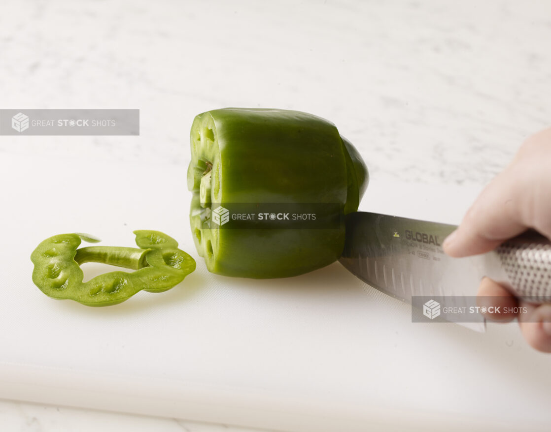 Whole green bell pepper, one end sliced off with a hand holding a kitchen knife