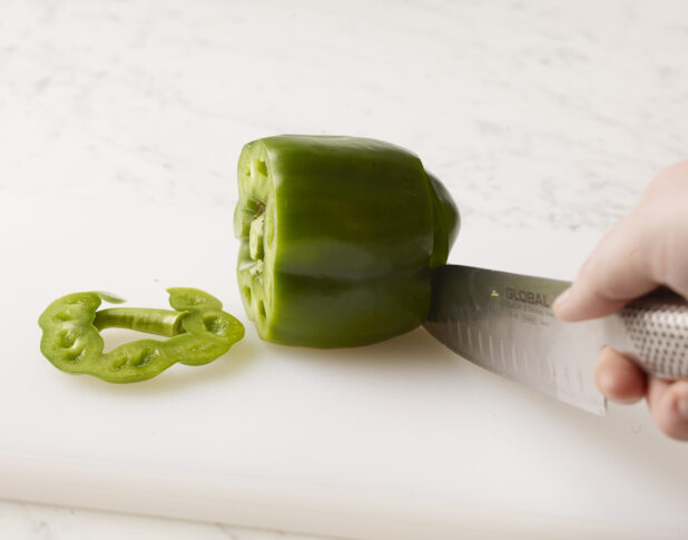 Whole green bell pepper, one end sliced off with a hand holding a kitchen knife