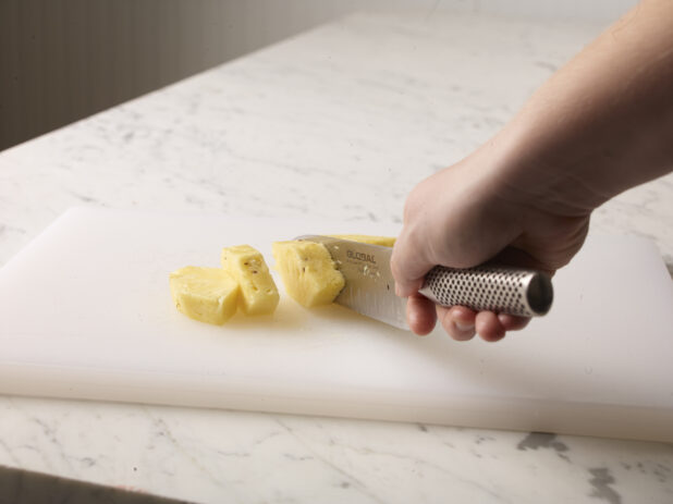 Hand cutting a cleaned pineapple on a white cutting board using a kitchen knife