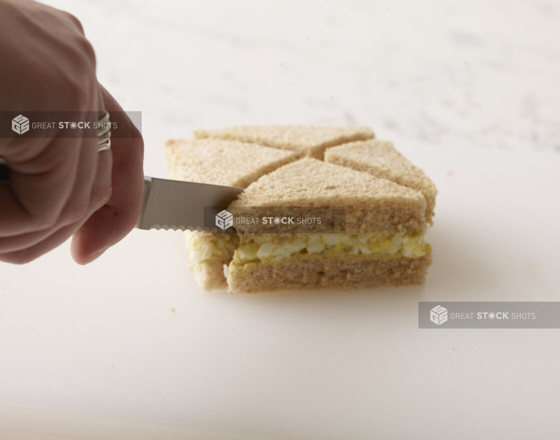 Hand cutting an egg salad finger sandwich with a serrated pairing knife on a white background