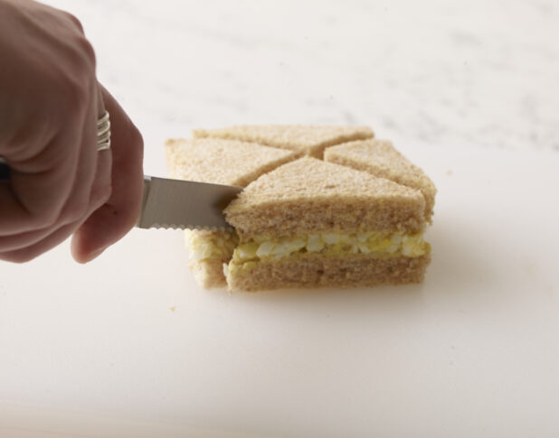 Hand cutting an egg salad finger sandwich with a serrated pairing knife on a white background