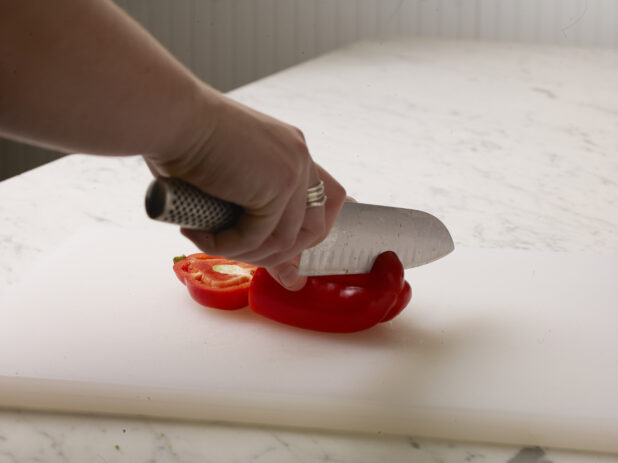 Woman cutting a red pepper on a white cutting board with a white marble background