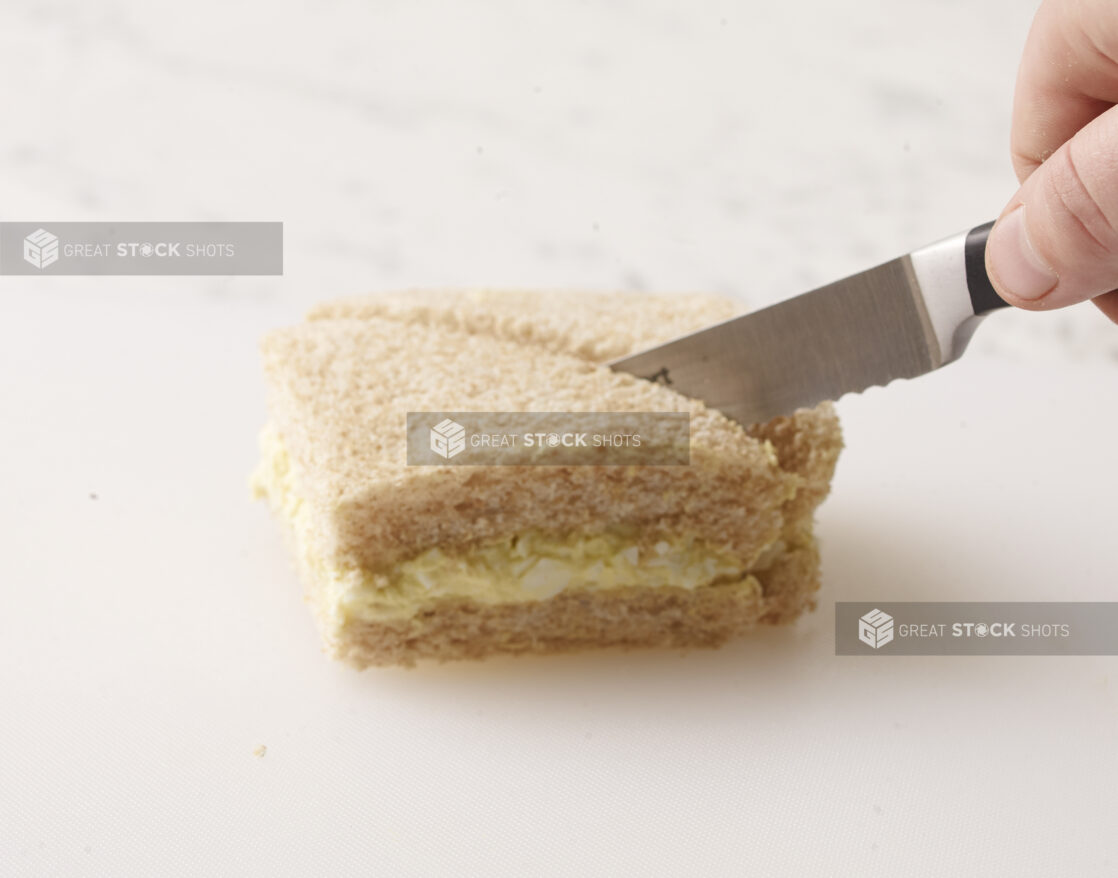 Hand cutting an egg salad finger sandwich in half with a serrated knife on a white background