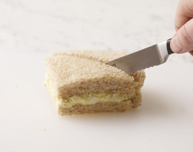 Hand cutting an egg salad finger sandwich in half with a serrated knife on a white background