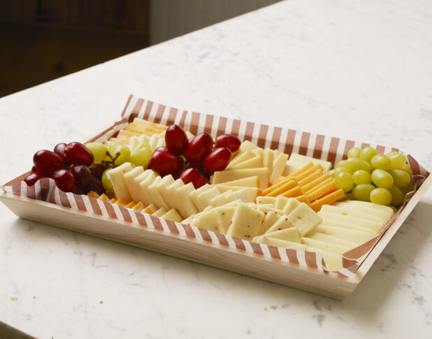 Assorted Block Cheeses Sliced and Arranged with Red and Green Grapes on a Wooden Serving Tray for Catering, on a White Marble Counter Top in a Kitchen Setting