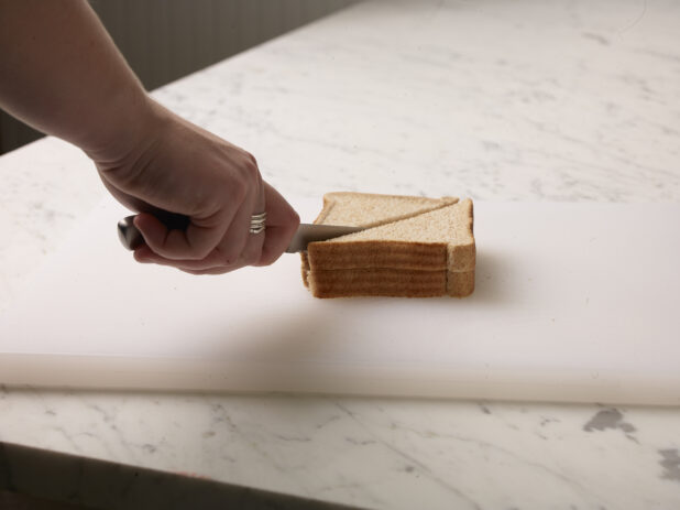 Woman cutting a sandwich in half on a white cutting board with a white marble background