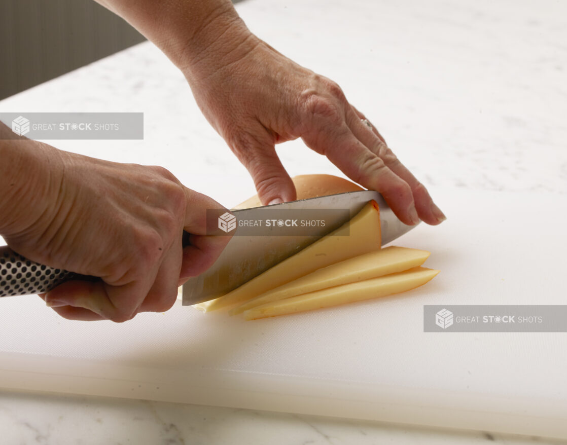 Woman cutting a brick of cheese into triangle slices on a white cutting board with a white marble background