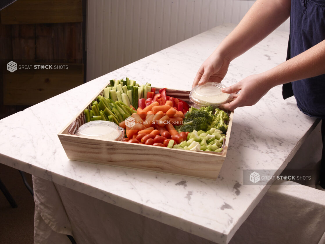 Person placing a lid on a dip inside of an assorted vegetable tray on a white marble countertop