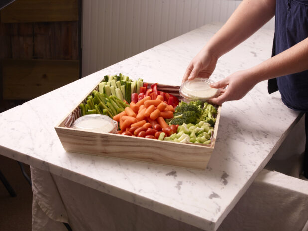 Person placing a lid on a dip inside of an assorted vegetable tray on a white marble countertop