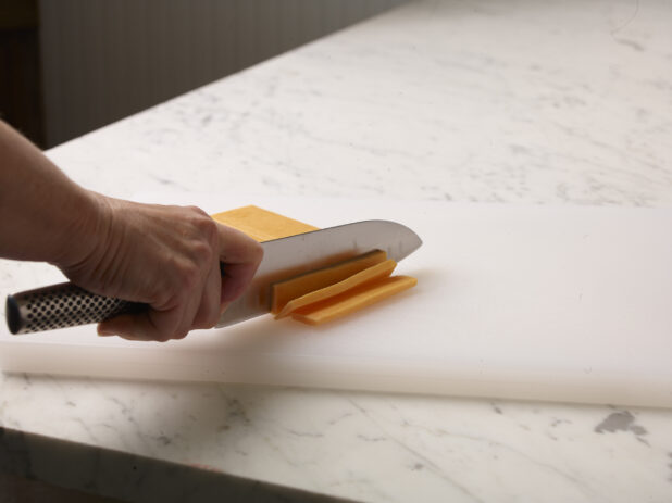 Woman cutting a brick of cheddar cheese into slices on a white cutting board with a white marble background