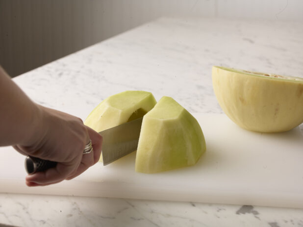 Hand using a knife to cut a honeydew melon on a white cutting board on a white marble background