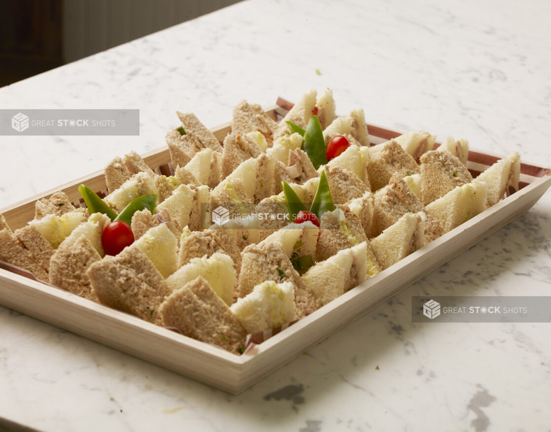 Wood catering tray lined with parchment paper filled with tuna and egg salad sandwich triangles on a marble countertop