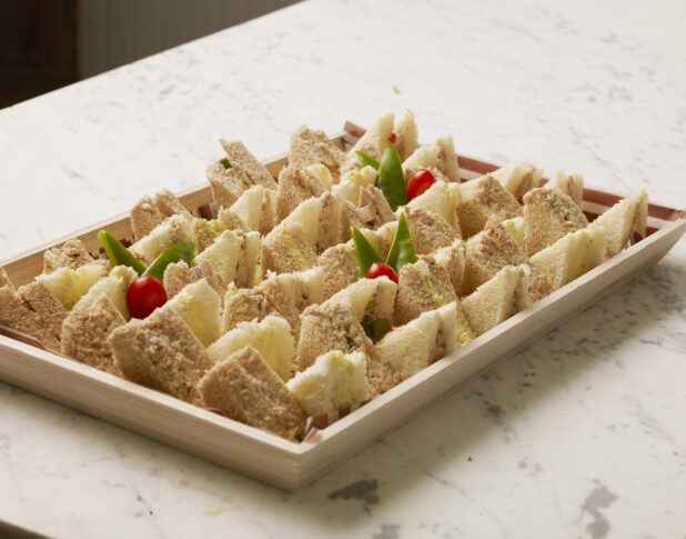 Wood catering tray lined with parchment paper filled with tuna and egg salad sandwich triangles on a marble countertop